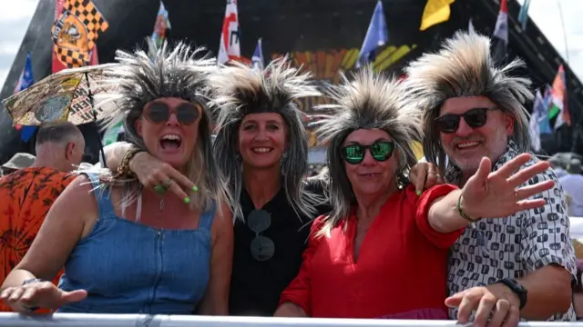 Four fans wearing wigs stand in front of the Pyramid Stage