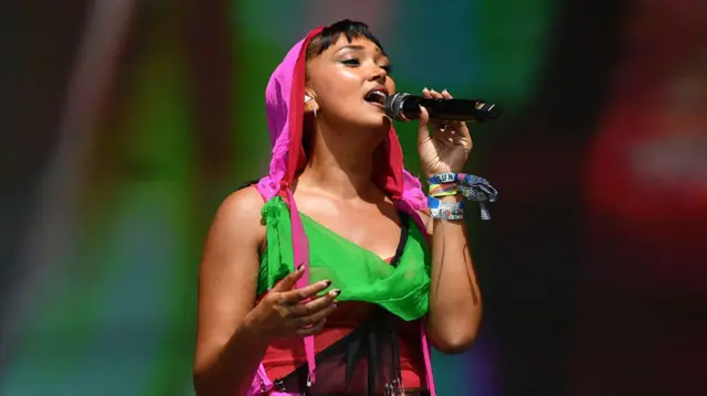 She is holding a mic in her left hand and wearing a bright pink and green outfit