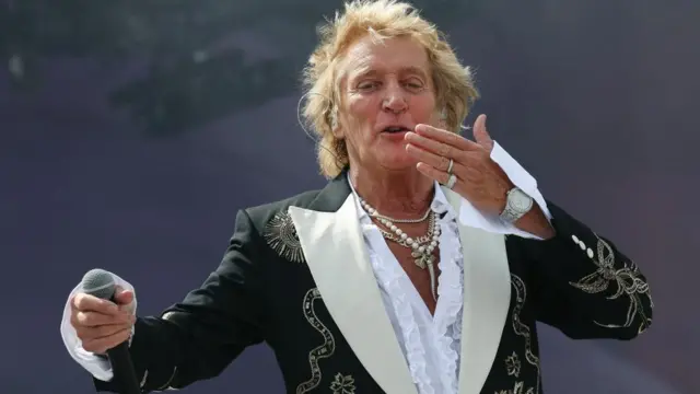 Rod Stewart performs on the Pyramid Stage - he's blowing a kiss and has a microphone in his other hand. He's wearing a black jacket with gold details and white lapels.