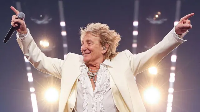 Rod Stewart smiling on stage wearing all white, his hands in the air, holding a microphone