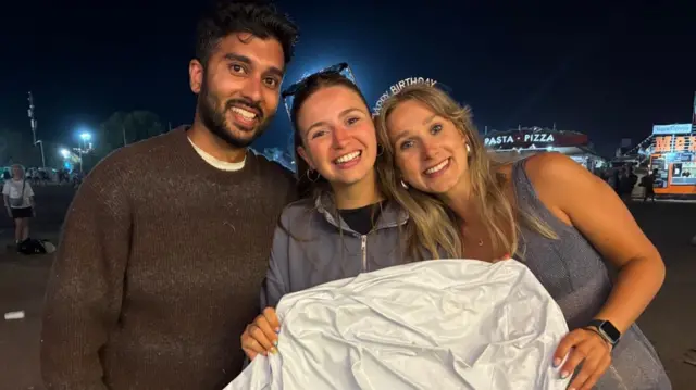 Three fans at Glastonbury pose for a photo holding a deflated white balloon, with some food stalls lit up behind them. A man with dark hair and a brown jumper smiles on the left, with a brunette girl in a blue-grey zip-up in the centre and a blonde girl on the right wearing a smart watch and a 'Happy Birthday' headband.
