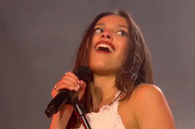 Olivia Rodrigo smiles while performing on stage - she is wearing a white dress and holding her microphone