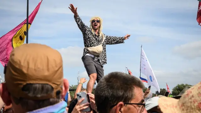 A man stands on up high above the crowd, wearing a Rod Stewart style wig