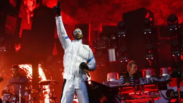 The backdrop has a red glow. Maxim is out front wearing all white and white paint over his eyes. He is holding a mic in his left hand a has his right arm up in the air and is pointing up with his index finger. His bandmates are behind on the drums and keyboard