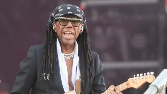 Nile Rodgers wearing black printer bandana, a black blazer and open white shirt underneath it. he has 3 strings of white beaded necklances, and sunglasses on
