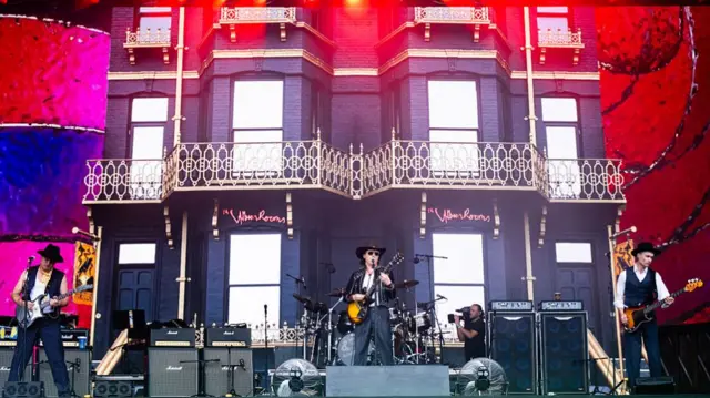 three members standing spaced out across the stage (left, centre and right), each with a guitar. The backdrop is a townhouse with a balcony.