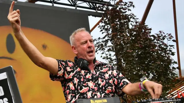 He's DJ'ing with his right index finger pointing up in the air. On his right is a sign that reads Drop acid not bombs