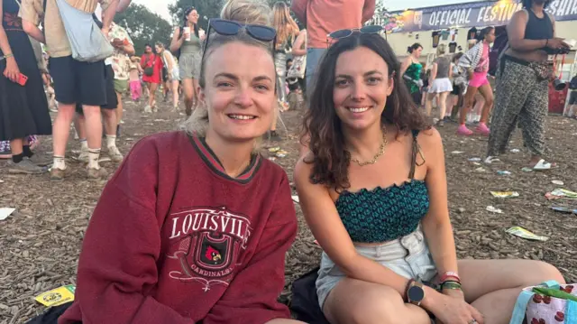 Two friends are sat cross-legged on the ground. The woman on the left has blone hair and is wearing a red jumper. The woman on the right is has brunette hair and it wearing a black and green crop top.