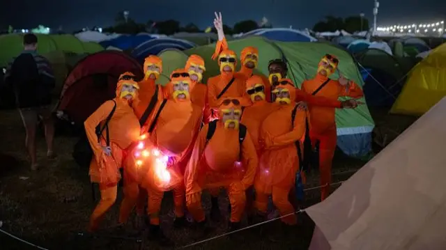 Several festivalgoers dressed as the Lorax, a character created by author Dr. Seuss. They're wearing orange jumpsuits and yellow moustaches and eyebrows and are gathered together a group photo. There are tents in the background.