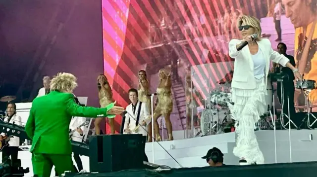 Rod Stewart in green at the foot of the stage, while Lulu wearing white with tasselled trousers performs on stage