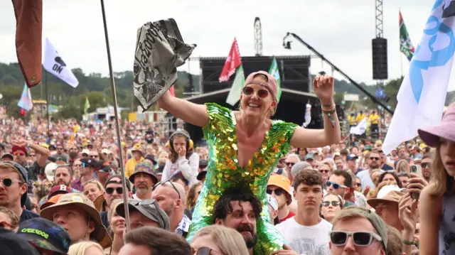 A woman wearing a green and yellow sequin dress cheers with her arms in the air as she sits on a man's shoulders in the crowd. There are people and flags all around her.