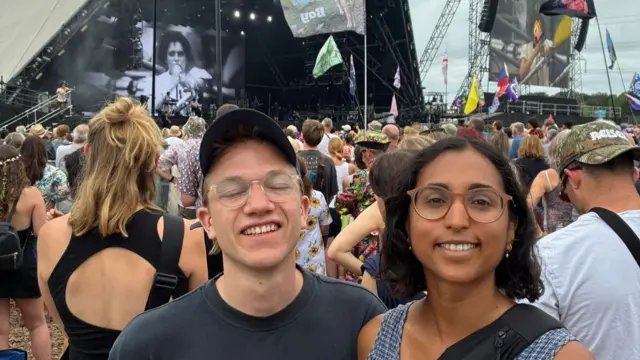 A man wearing a hat and a woman smiling, in front of the Pyramid Stage.