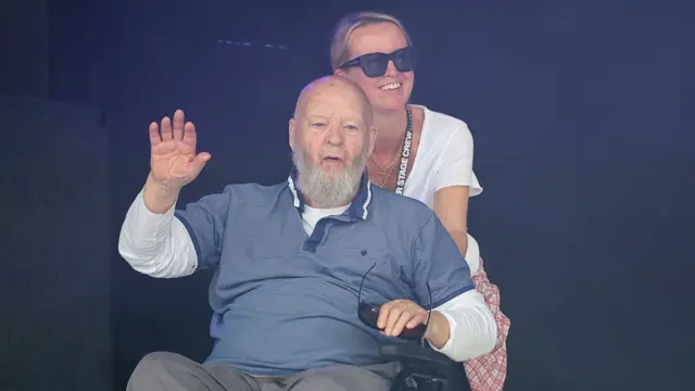 Sir Michael Eavis sitting in a wheelchair pushed by his daughter Emily Eavis