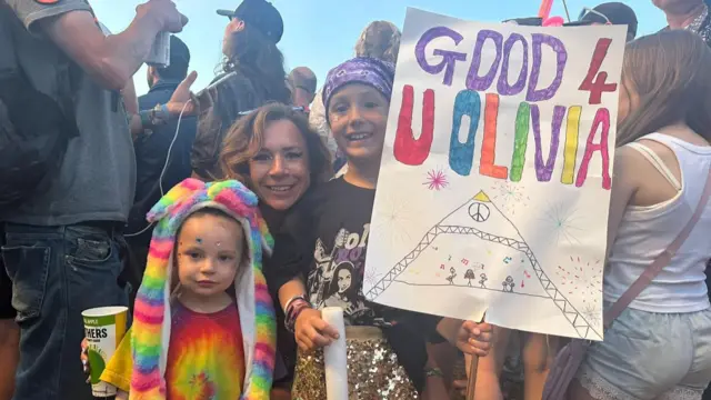 Two children and their mum with a sign reading good 4 u Olivia