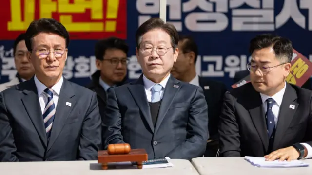 Kim and Lee, dressed in suits, sit in front a desk at a rally