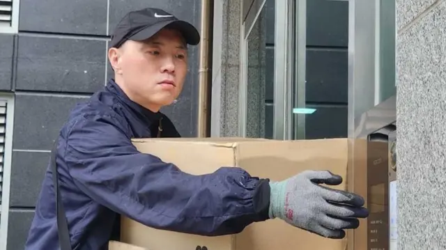 Cho Shin-hwan, a South Korean delivery worker, holds a cardboard package as he buzzes the door of an apartment building.