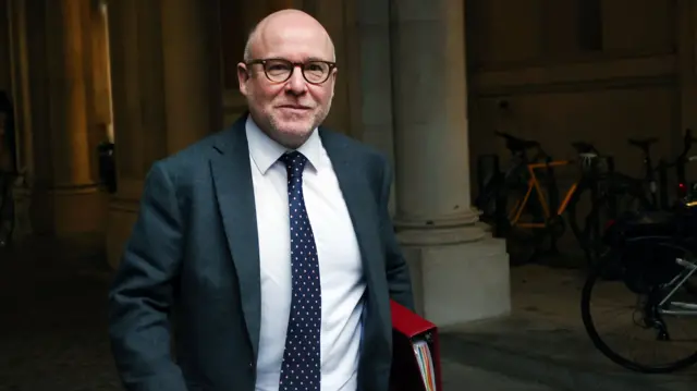 Richard Hermer, wearing a suit and holding a red binder, walks on a street