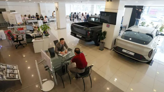 A wide shot of a car dealership shows people queuing to vote in the background.