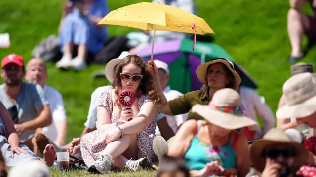 People try to keep cool with a fan and umbrella on the grass at Wimbledon