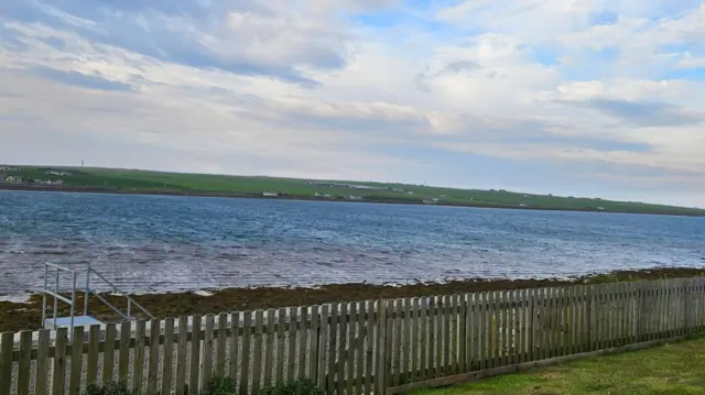 A body of water splits some land with a fence on one side and green small hills on the other.