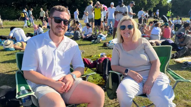 Debbie and Reece sit on some deck chairs with the backdrop of those in the queue at wimbledon