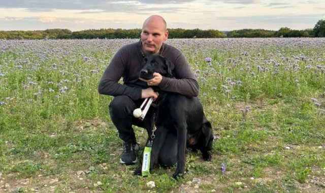 Sean Dilley and his guide dog, a black Labrador, in a field of flowers