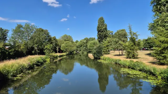 A river flowing ion between fields, there are very few clouds in the very blue sky.
