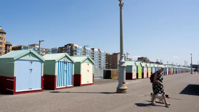 Sunny weather on beachfront, beach huts in a line, multicoloured, with a woman walking a dog nearby.