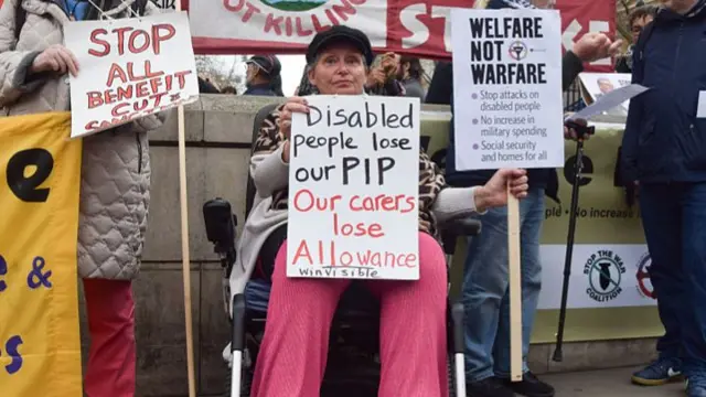 A woman in a wheelchair holds a protest sign that reads disabled people lose our pip our carers lose allowance