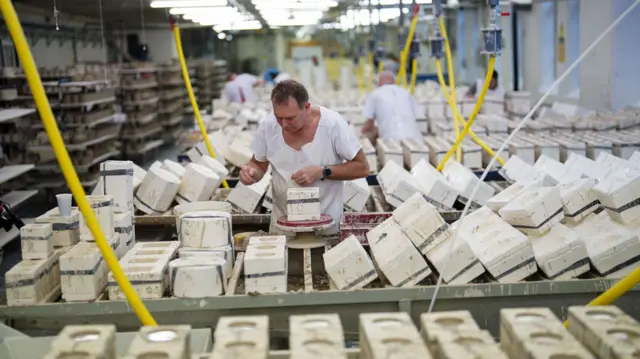 A man wearing an all-white outfit, stands working at a ceramics production line in a factory. More workers can be seen behind him and on the left are shelves full of finished pieces.