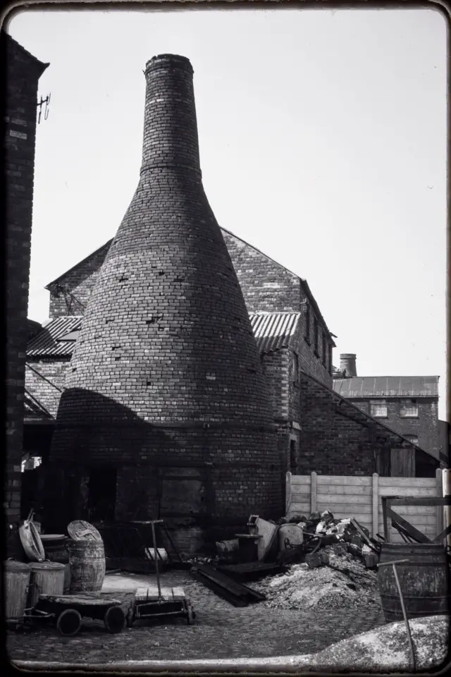 A black and white image in a pottery yard at the bottom of a bottle kiln with buildings behind it. The yard has hand-pulled trailers and piles of rubble as well as barrels and tools.