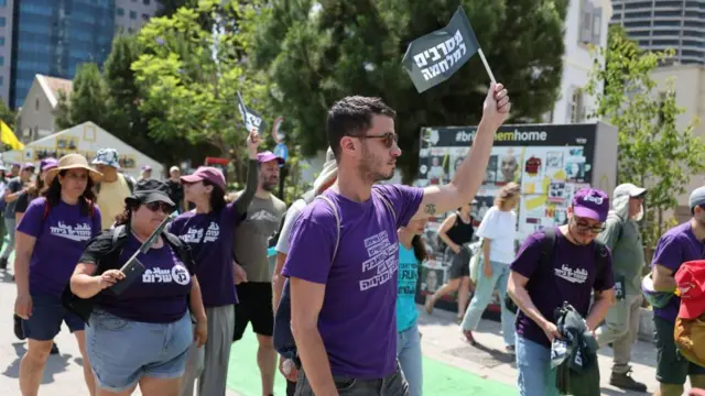 Israeli activists wearing purple T-shirts. In the centre of the photo is a man holding up a black flag with Hebrew writing.
