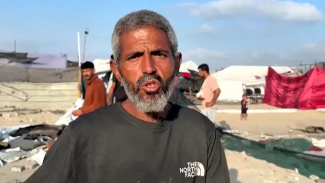 A man speaking as people walk behind him on a dirt road with tents littered about