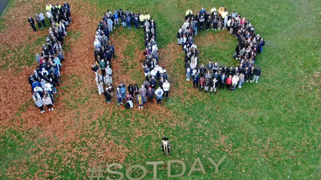An aerial image of a group of people who have formed the number 100. In front of them is a figure dressed in mayoral attire who is next to a display of large letters that say "#SOTDAY".