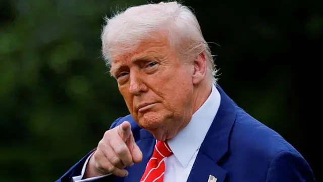 Donald Trump, wearing a blue suit and red tie, points to the camera as he walks on a lawn outside the White House