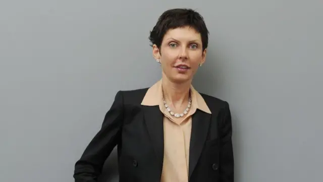 A woman with short dark hair, earrings, a necklace, a peach-coloured shirt and a black jacket, stands facing the camera in front of a grey wall.