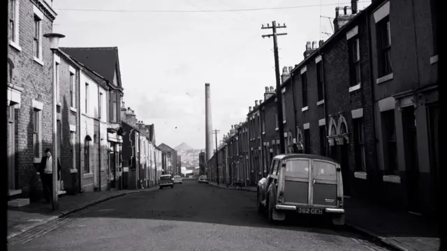 An old black and white image of a residential street, with terraced houses on either side. Old cars are parked at the side of the road and a man is walking towards us on the left. A large chimney tower is visible in the distance.