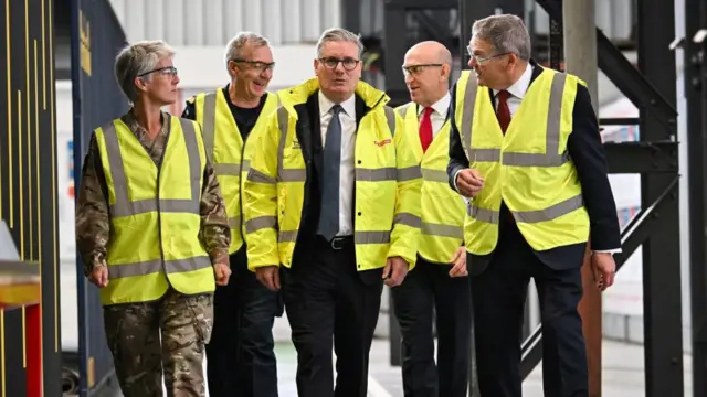 Starmer and defence secretary Healey are wearing high vis vests and walking through a job site. they're with other military staffers, including one dressed in fatigues.