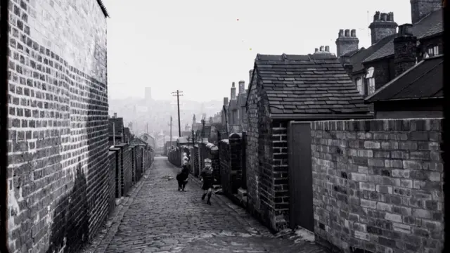 A black and white image of an alleyway between two rows of terraced houses. A boy is running towards the camera with a dog behind him.