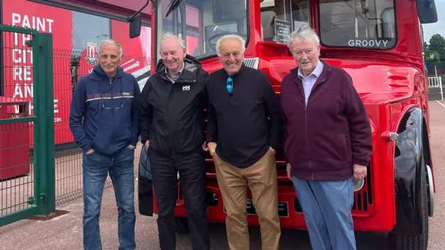 Four elderly men stand in a line in front of a red bus.