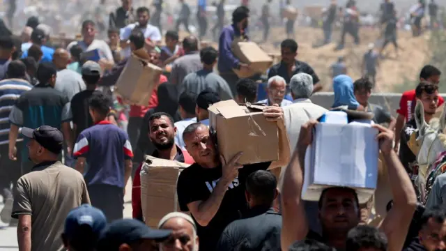 Dozens of Palestinian men can be seen carrying cardboard boxes believed to contain aid from GHF. They're carrying them on their head or shoulders. It is a sunny day and some are squinting.