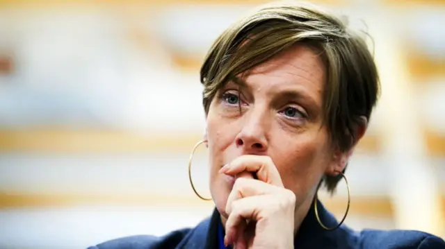 Jess Phillips (woman in her 40s with short brown hair, golden hoops) wearing a blazer as she looks ahead with her left hand over her mouth