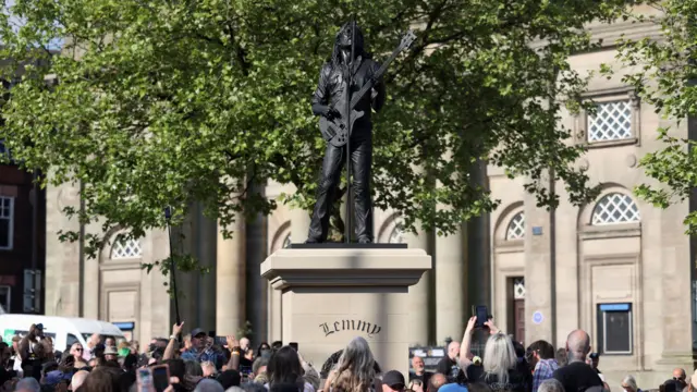 A crowd, some of them holding up phones, gathers around a beige-coloured plinth which has a black statue on top of Lemmy - a man with long hair holding a bass guitar and singing upwards into a microphone. A large building can be seen in the background.