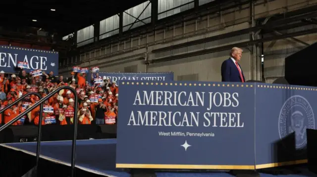 Trump stands at a podium behind a sign reading 'American Jobs, American Steel' - West Mifflin Pennsylvania. A crowd of people wearing high-vis and hard hats stand behind him