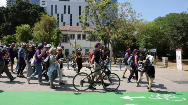 Israeli activists walking, with many wearing purple T-shirts. In the centre of the photo is a man walking with his bike.