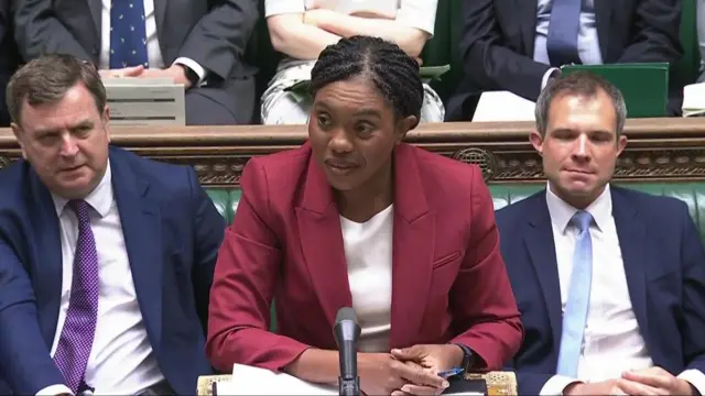 Kemi Badenoch wears a wine red suit and stands in the House of Commons.