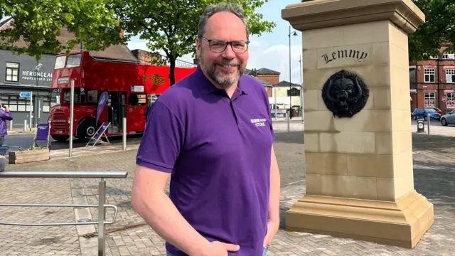 A man wearing a purple BBC Radio Stoke T-shirt, stands with his hands in his pocket on a market square, in front of the statue of Lemmy Kilmister