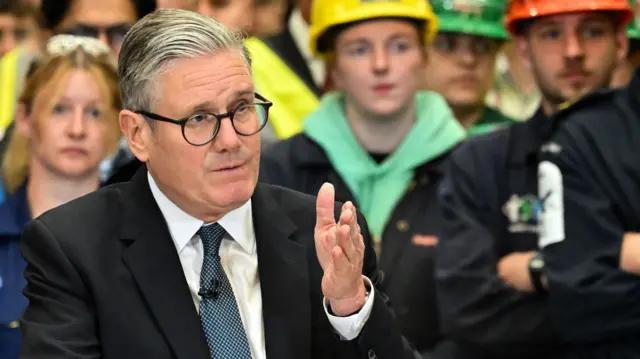 Prime Minister Sir Keir Starmer speaks during a visit to BAE Systems in Govan, Glasgow, to launch the Strategic Defence Review. He is in a black suit jacket with patterned blue tie. Blurred in the background is a group of people in hard hats.