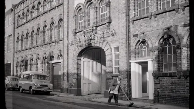 A black an white image of a large three-storey building with rows of windows and a large, arched entrance. Above the entrance it says Queen Anne China. A man is sweeping the pavement and two cars are parked at the side of the street.