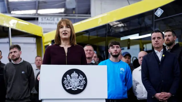 Rachel Reeves in burgundy zip-up blazer stands behind a white lectern bearing the symbol of the Treasury as she speaks inside a factory